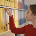 Side view of contemplating female assistant in casual style standing near shelves and choosing file with documents